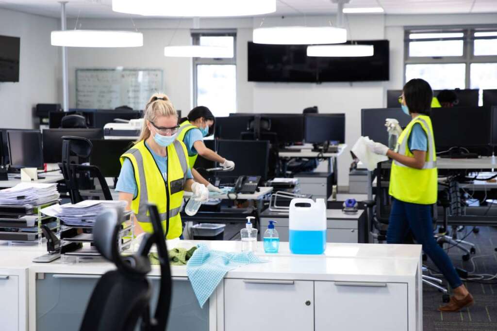 Caucasian woman cleaning an office wearing hi vis vest, gloves and face mask, using disinfectant, colleagues working in background. Hygiene in workplace during Coronavirus Covid 19 pandemic.