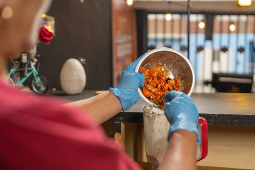 Cook is pouring a mixture of fried vegetables into an industrial blender, wearing gloves for food preparation