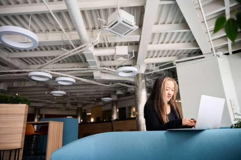 Focused asian female IT employee typing on laptop on sofa in coworking office