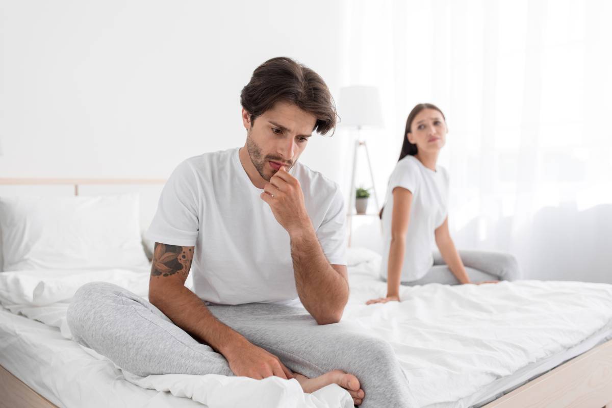 Depressed millennial european husband with stubble sits on bed and sad, wife apologizes in bedroom interior. Woman calm husband with erectile dysfunction, weakness and impotence. Sorry after quarrel